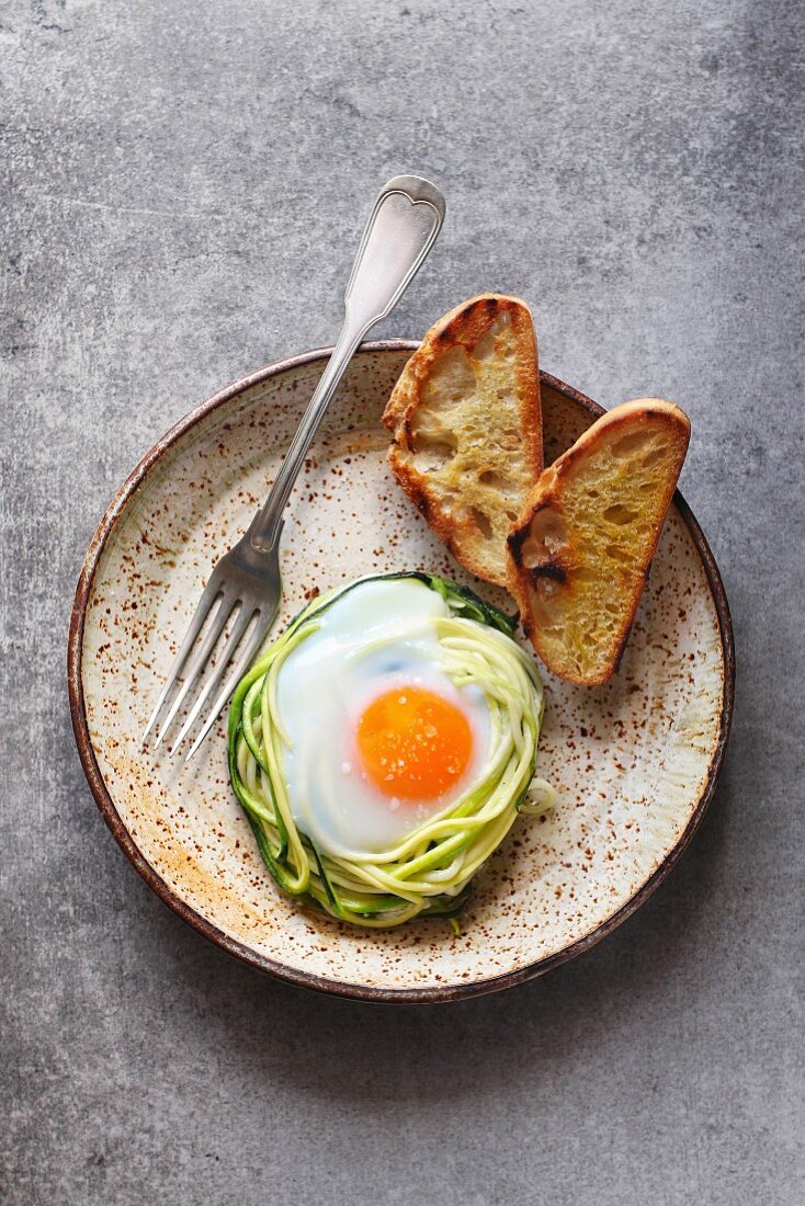 Breakfast with baked egg in zucchini noodles nest with bread toast on a ceramic plate