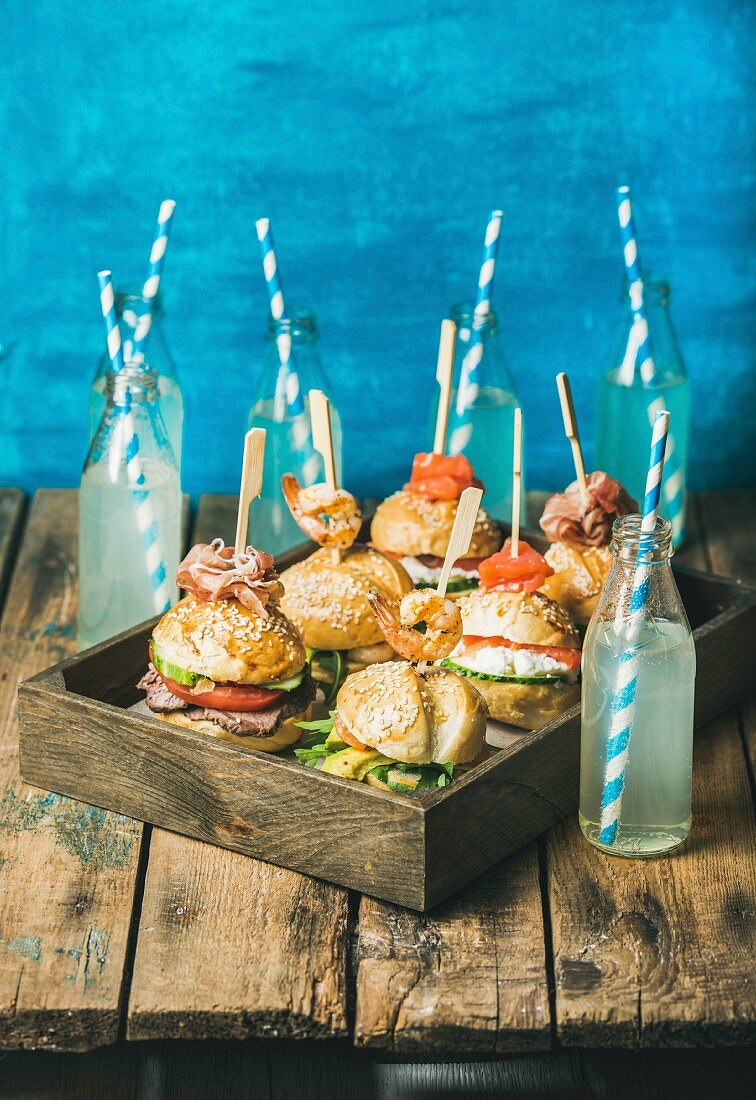 Different burgers with sticks in wooden tray and lemonade in bottles with straws on rustic shabby table