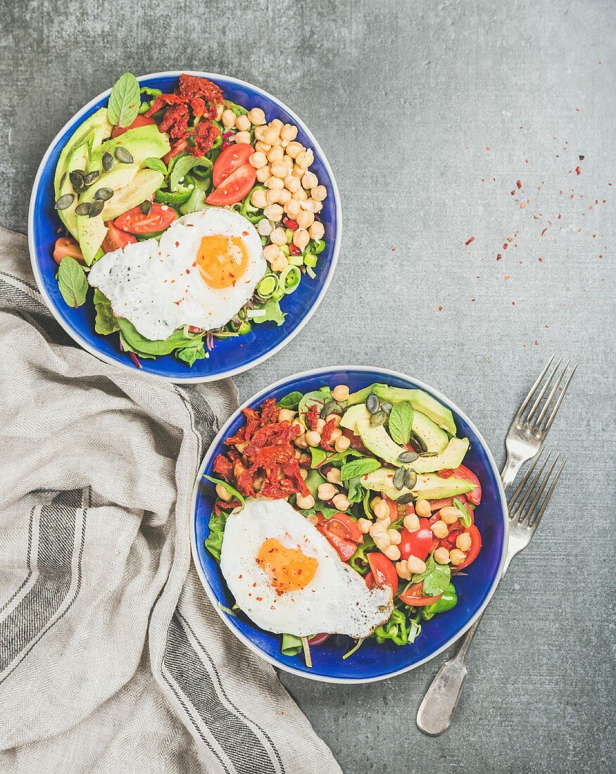 Healthy breakfast bowls with fried egg, chickpea sprouts, seeds, vegetables and greens over grey concrete background
