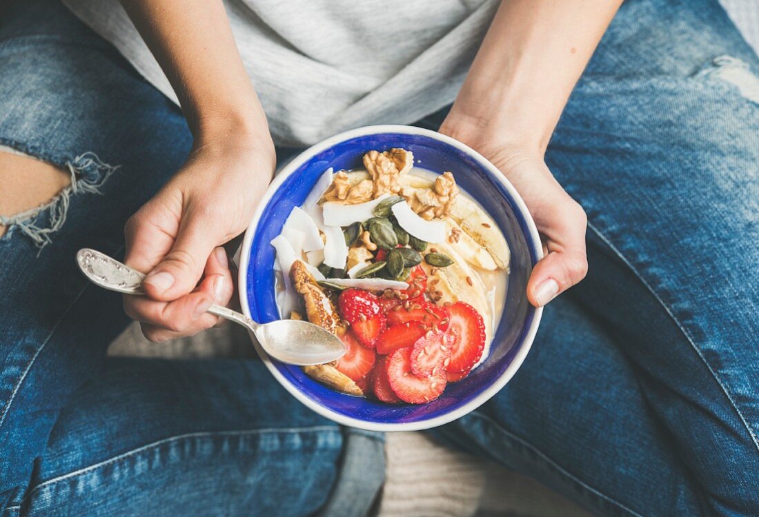 Yogurt, granola, seeds, fresh and dry fruits and honey in blue ceramic bowl in woman's hands