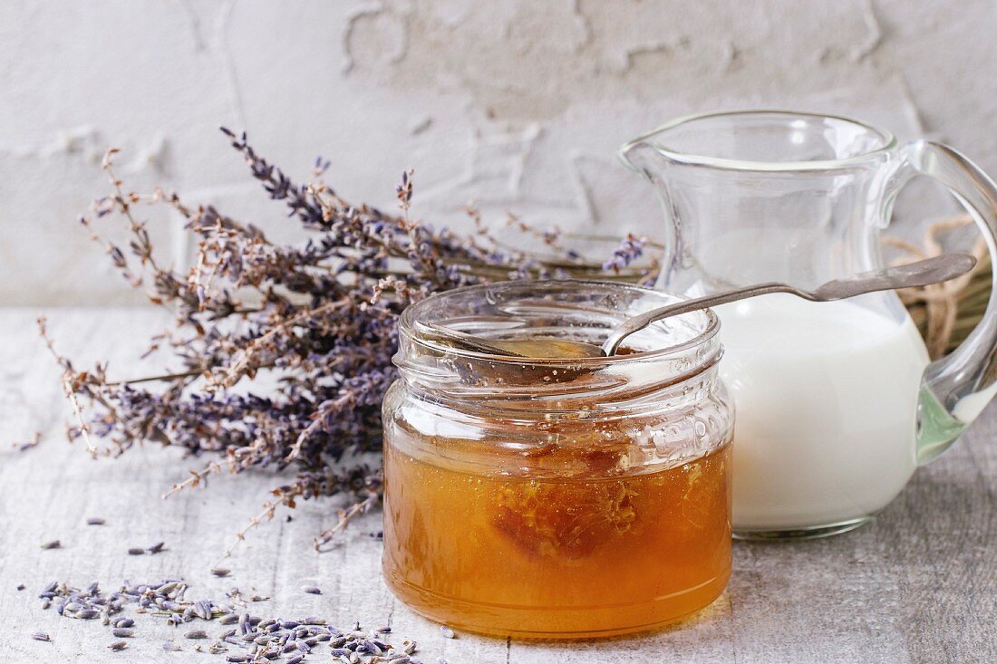 Geöffnetes Honigglas mit Honigwabe und Löffel, daneben Milchkrug und Lavendel