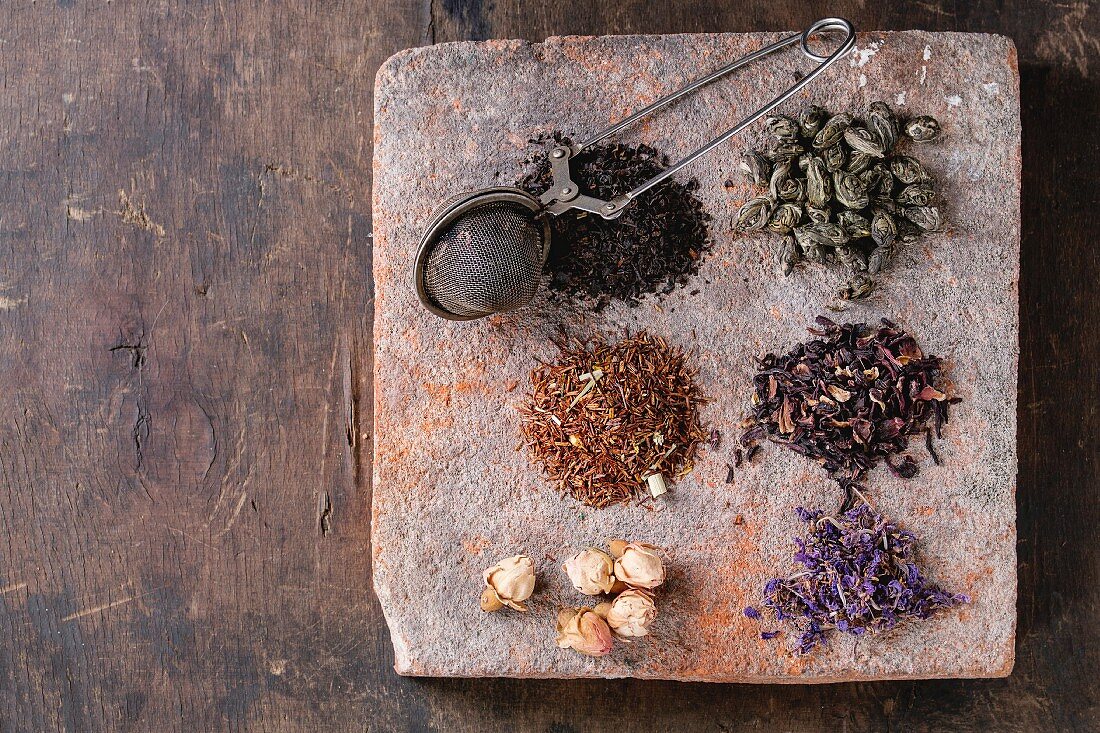 Variety of black, green, rooibos, herbal dry tea leaves and rose buds with vintage strainer on terracotta board