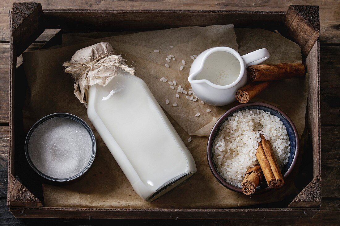 Ingredients for making rice pudding: Bowl of white uncooked rice, sugar, cinnamon sticks, milk and cream in wood box