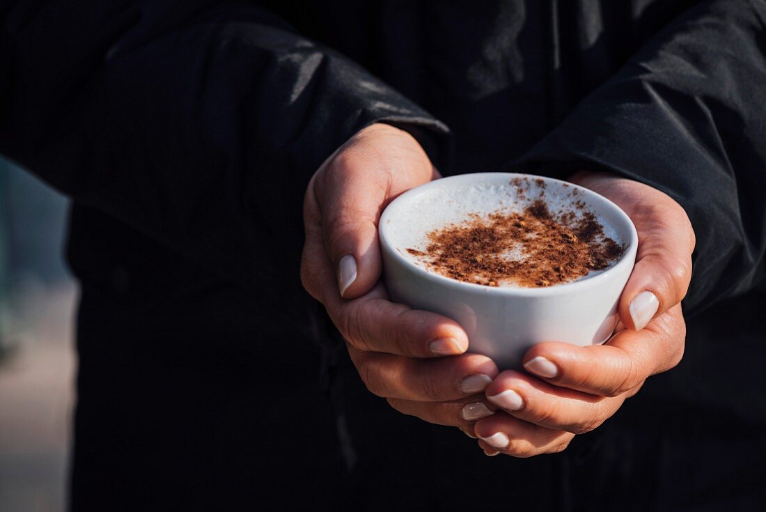 Hands holding a cup of Turkish sahlep topped with cinnamon on the street