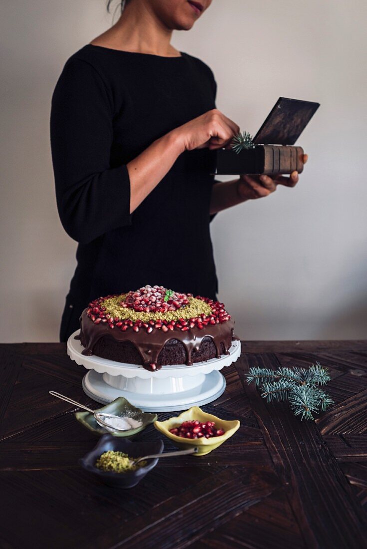A woman with a gift box, table with a Christmas Chocolate Cake and bowls with pomegranate seeds, pistachio and powdered sugar
