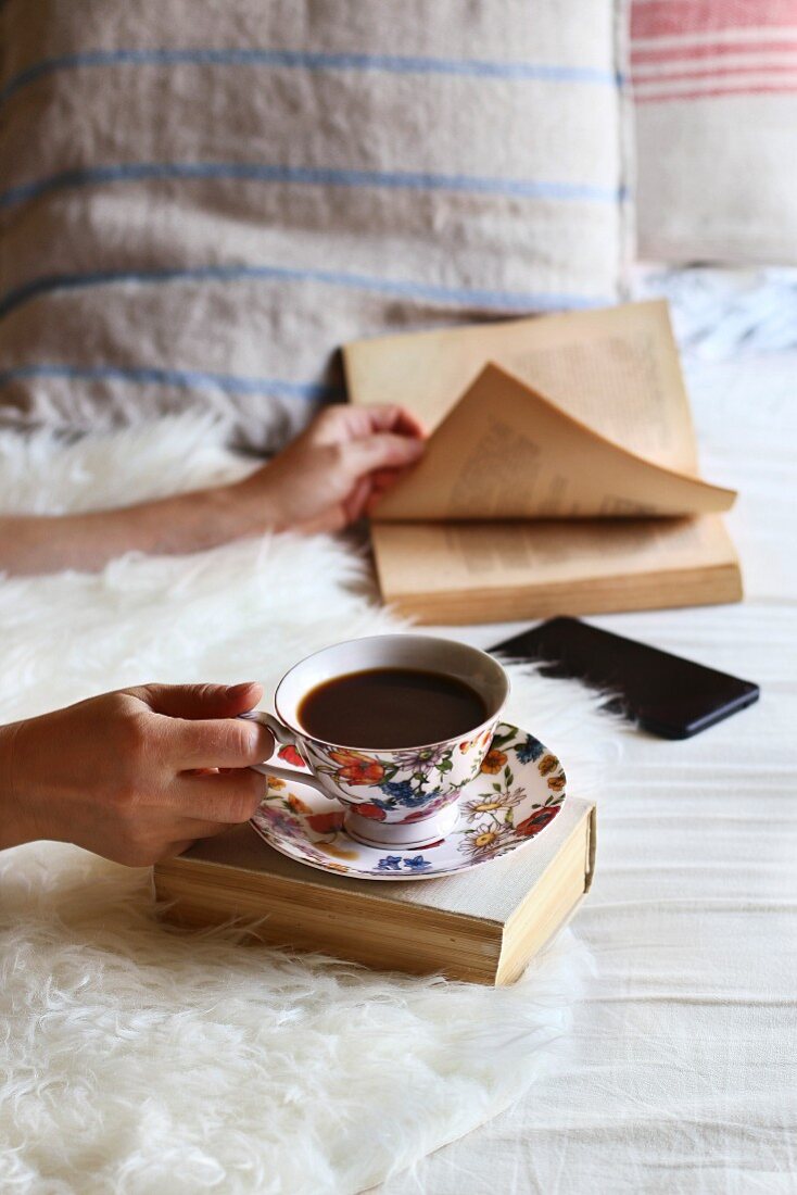 Female sitting on the bed, leafing through a book and drinking a cup of coffee