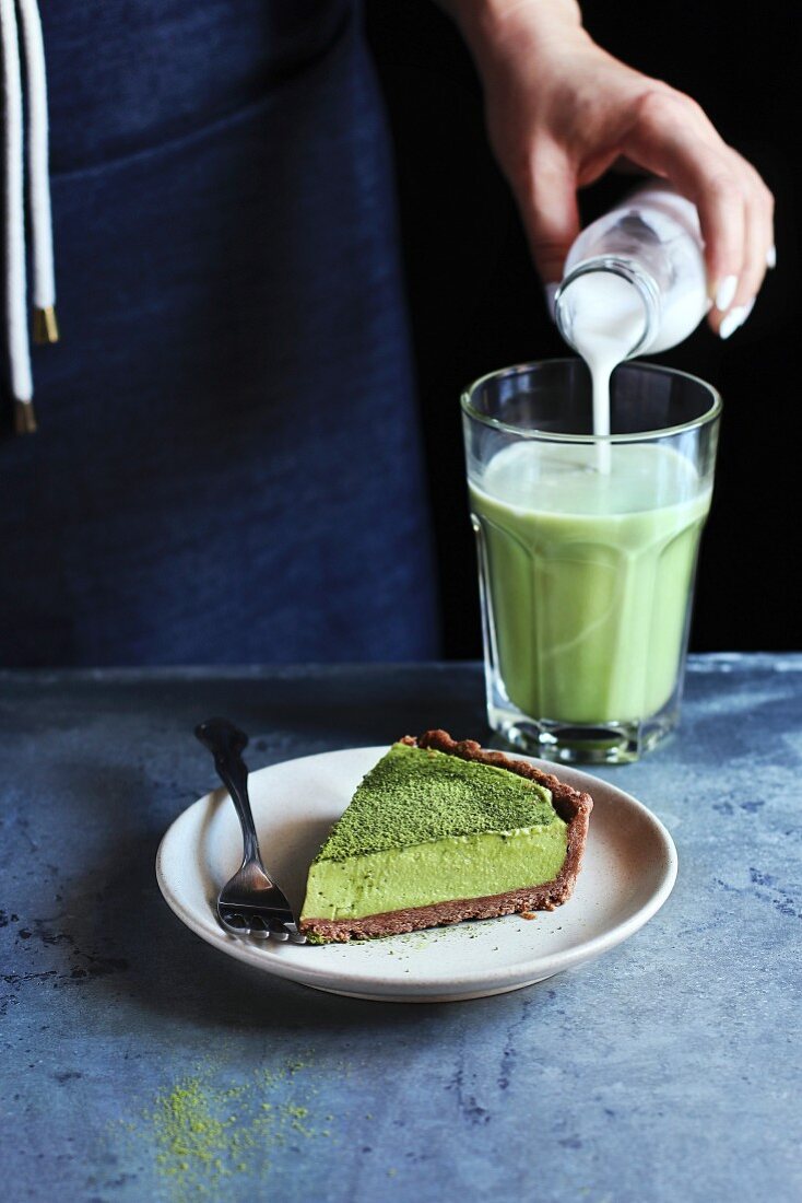 A slice of matcha tart with cream cheese filling on a plate, hand pouring coconut milk into matcha latte
