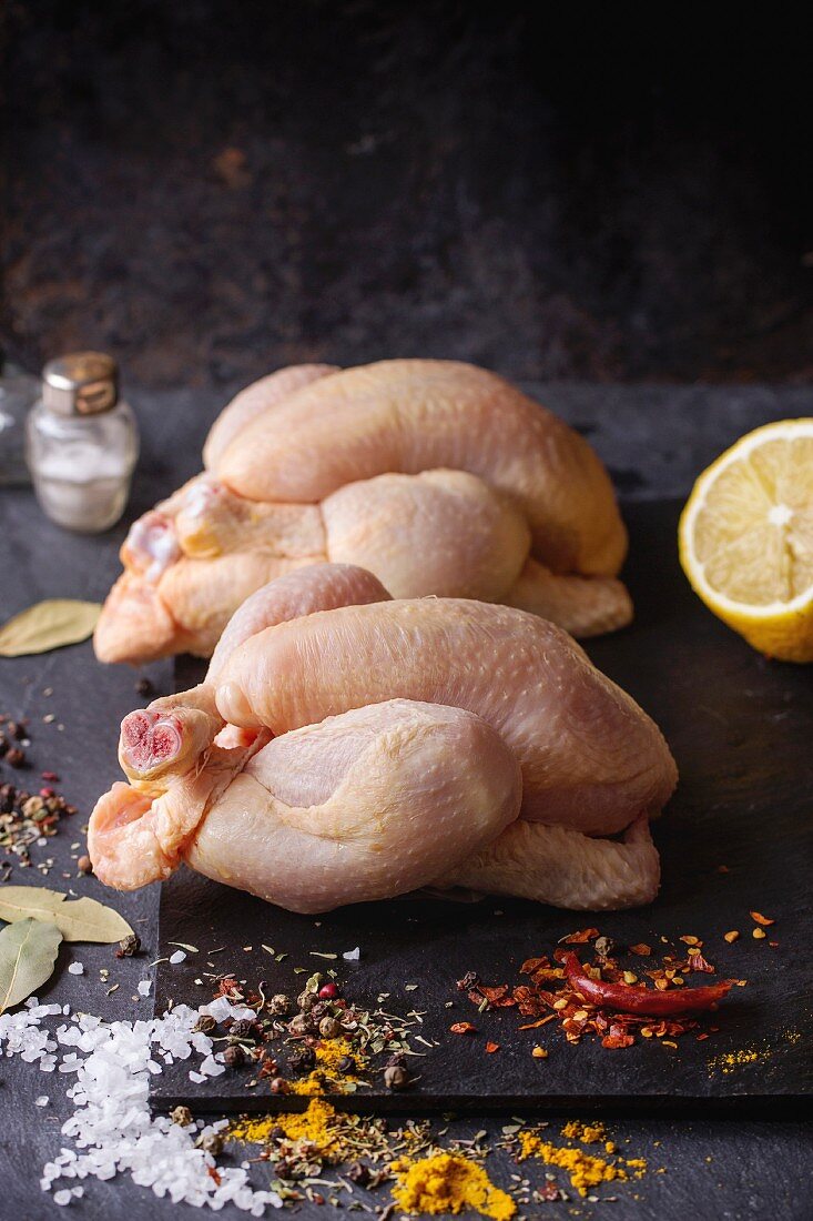 Two Raw mini Chicken with sea salt and different spices over black slate board