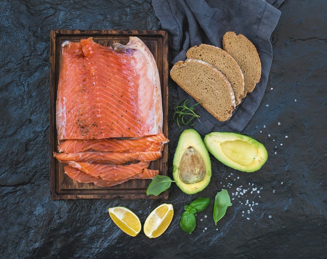 Smoked salmon filet with lemon, avocado, fresh herbs and bred on wooden serving board over dark stone backdrop