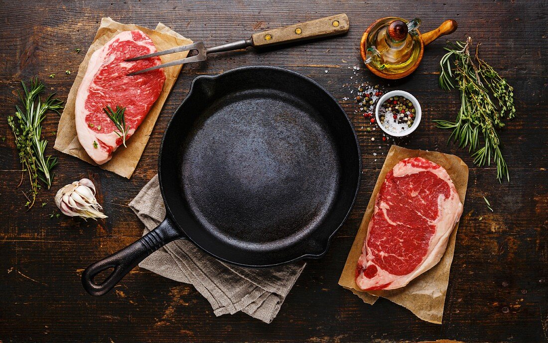 Bratpfanne umgeben von zwei rohen Striploin-Steaks, Kräutern und Gewürzen