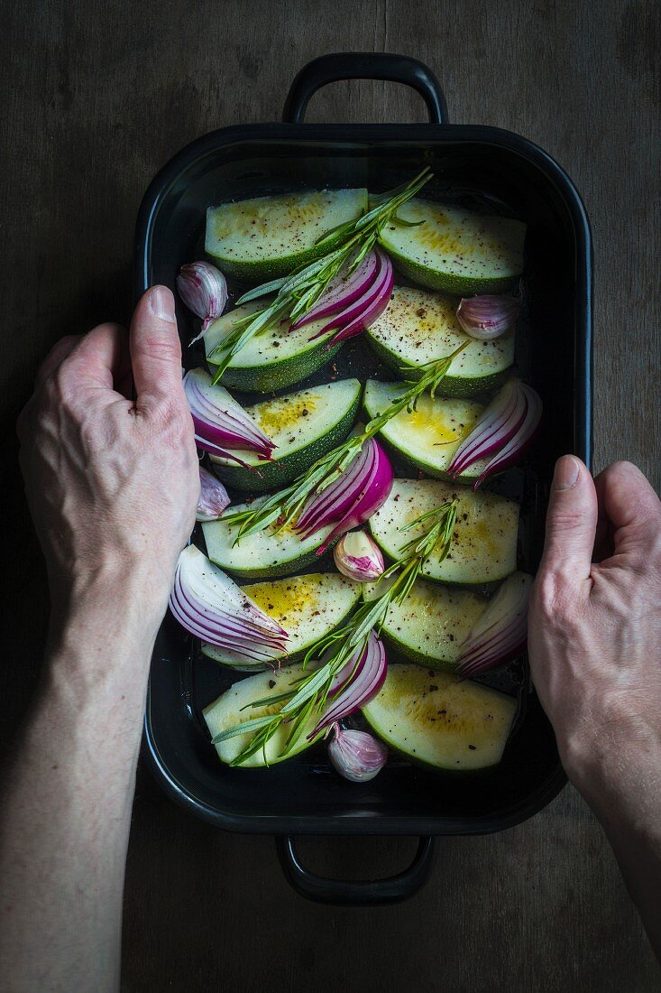 Hands holding an oven dish with courgette, onions and fresh herbs prepared for cooking