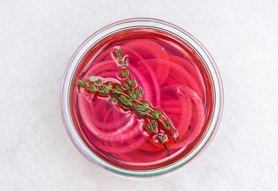 A jar of pickled red onions with thyme leaves is on a table