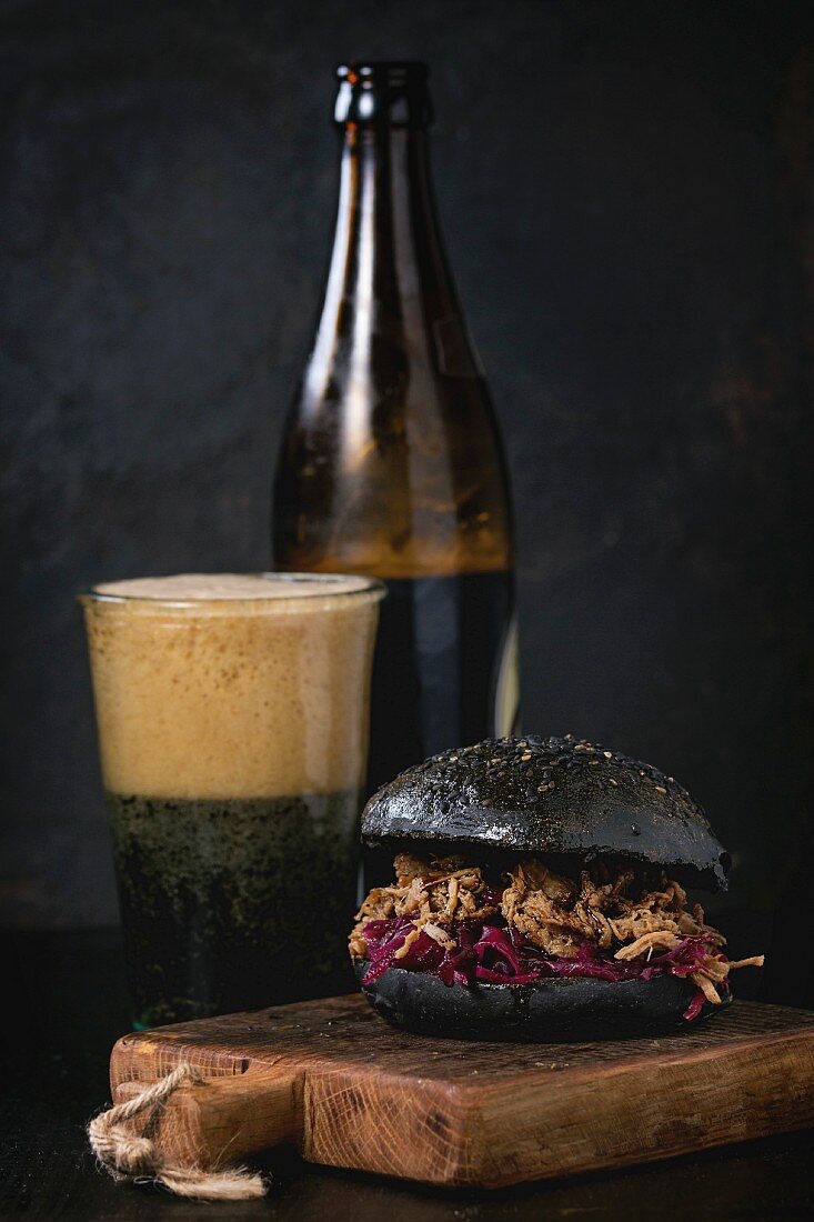 Black homemade burger with stews and red cabbage served on chopping board with glass and bottle of dark beer