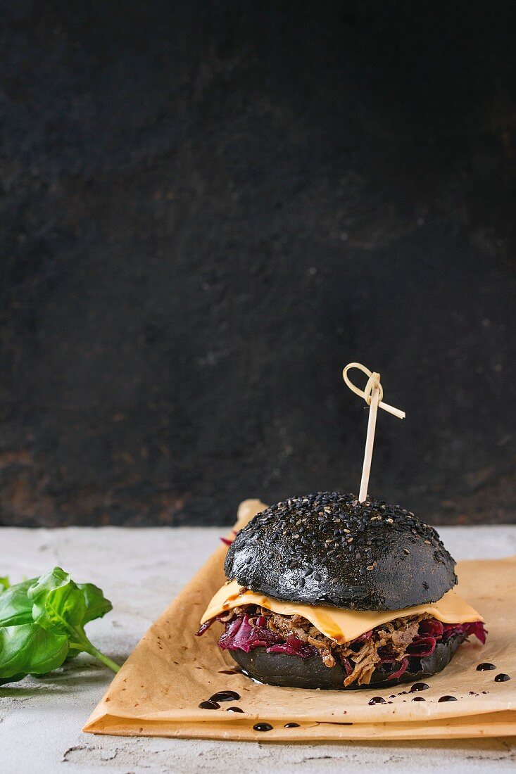 Black burger with beef stews, cheese, red cabbage and balsamic sauce served on baking paper with fresh basil