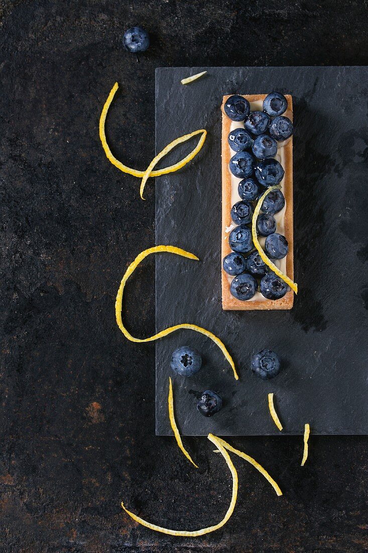 Square Lemon tartlet with fresh blueberries, served on black stone slate with lemon zest over black background