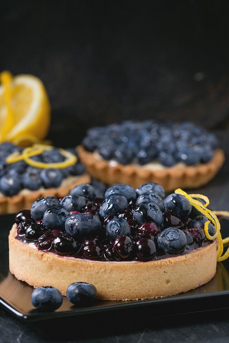 Lemon Tart and tartlets with fresh and cooked blueberries, served on black square plate with lemon and lemon zest