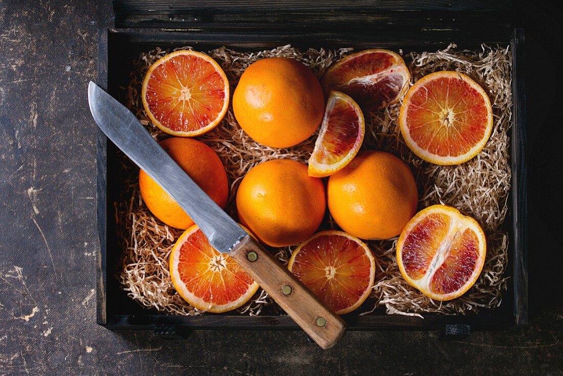 Sliced and whole Sicilian Blood oranges fruits in black wooden box with sawdust over dark background