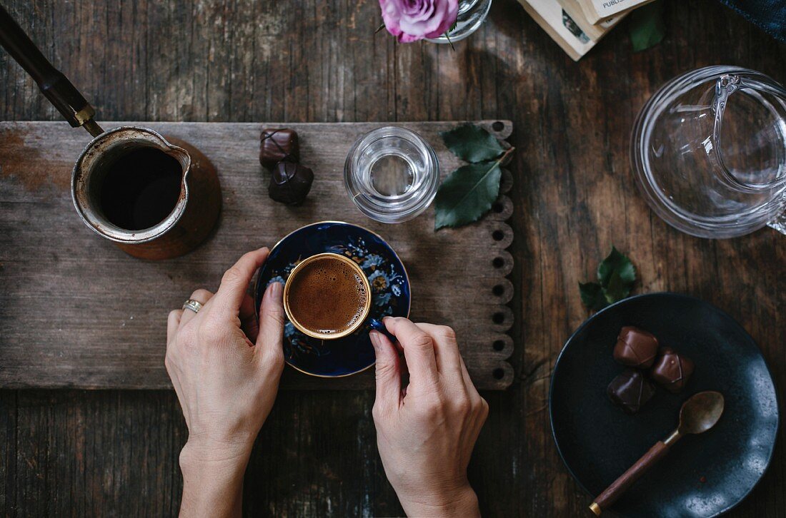 A woman is holding a cup of Turkish coffee with foam on top (with both hands) that is served with water and chocolate