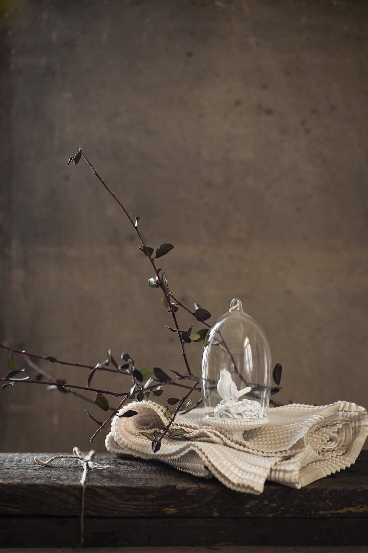 Glasglocke mit kleinem Porzellanvogel und Zweige auf weisser Tischdecke