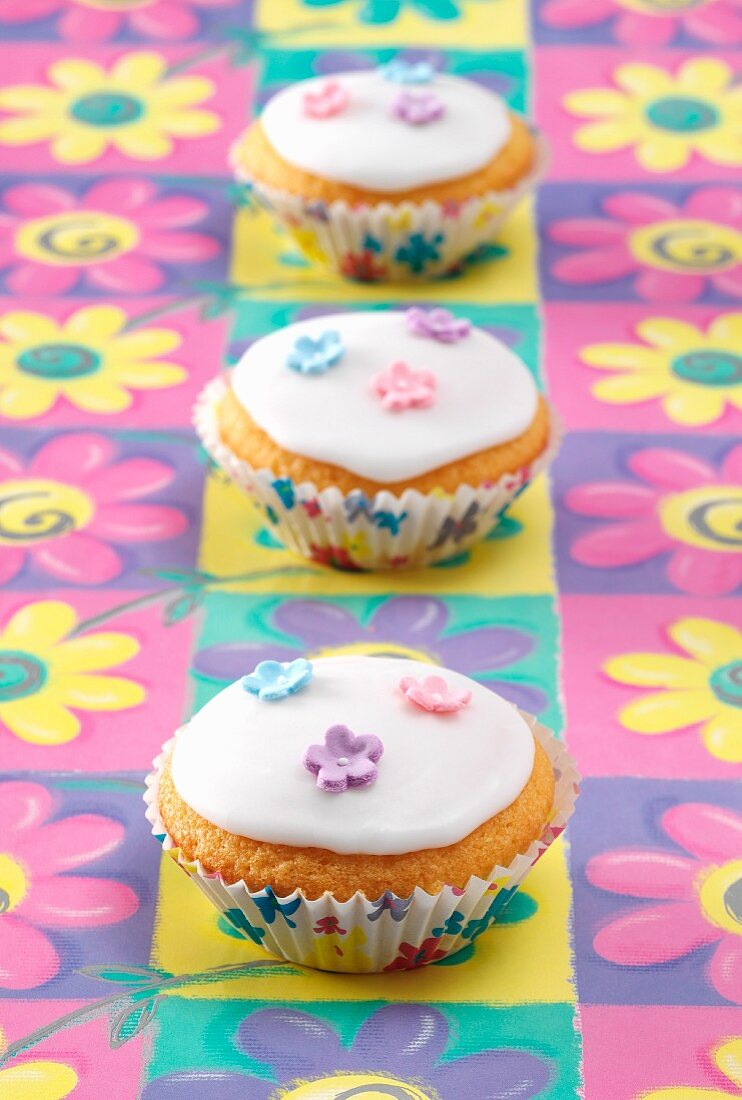 Three Fairy cakes sitting on a coloured flower pattern background