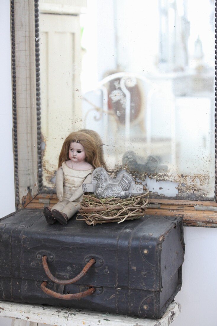 Doll next to chicken-shaped cake tin in nest on top of old suitcase