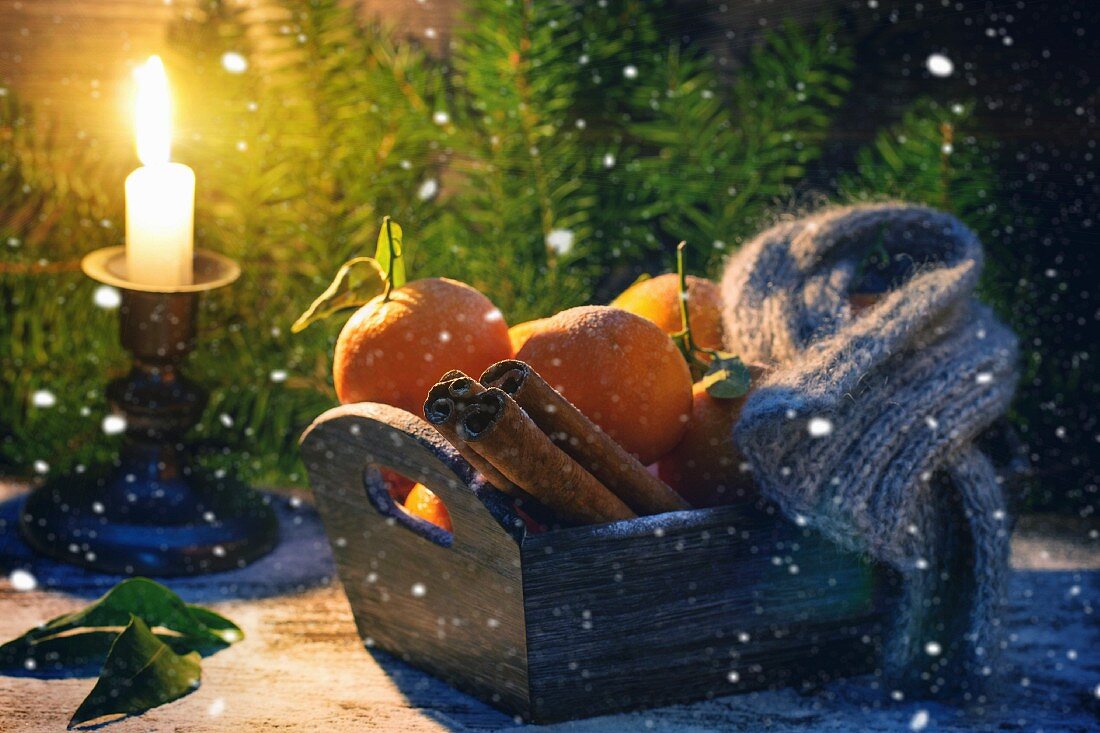 Mandarinen, Zimt und Schal in Holzkiste, dahinter brennende Kerze, Schnee und Weihnachtsbaum