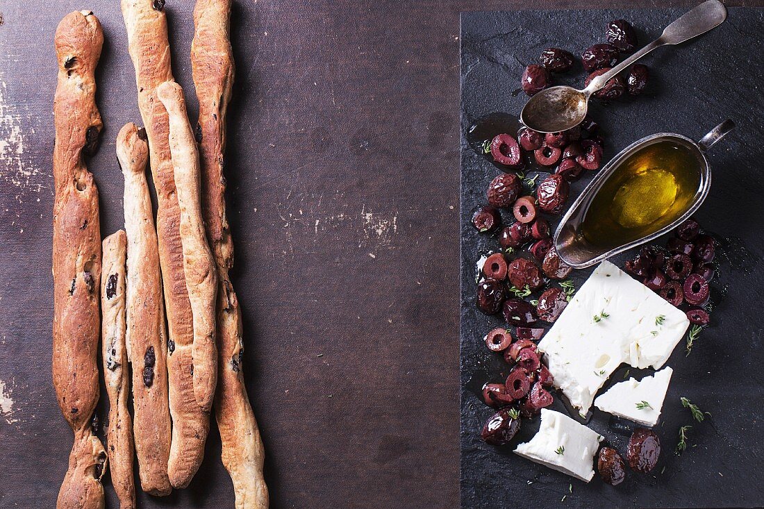 Whole and sliced black olives and block of feta cheese with olive oil and homemade grissini bread sticks