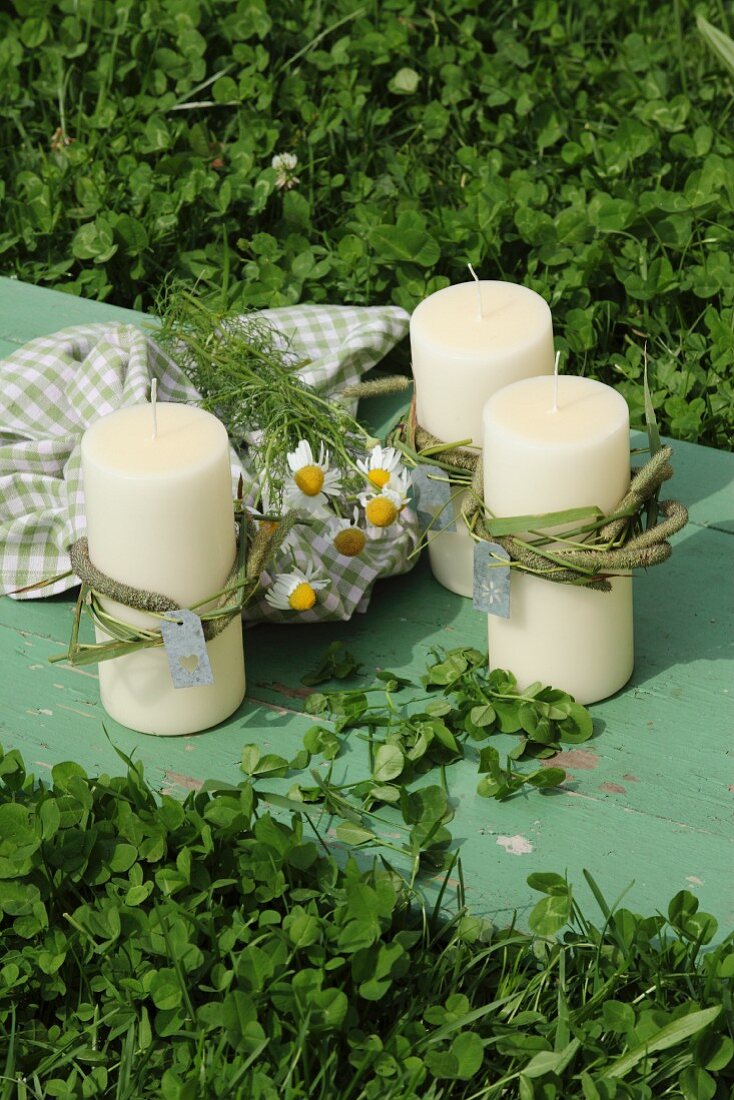 Candles decorated with chamomile flowers and checked tea towel on vintage wooden board amongst clover