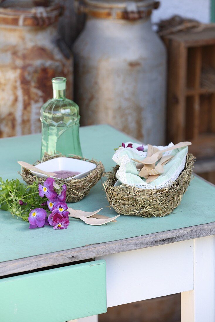 White porcelain bowls with wooden cutlery in hay baskets and horned violets on a vintage kitchen table