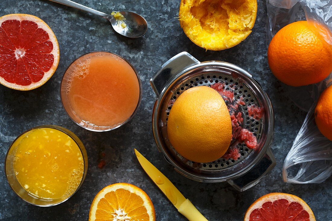 Manual metal citrus fruit extractor next to glasses of orange juice and red grapefruit juice and whole and half cut oranges and red grapefruit