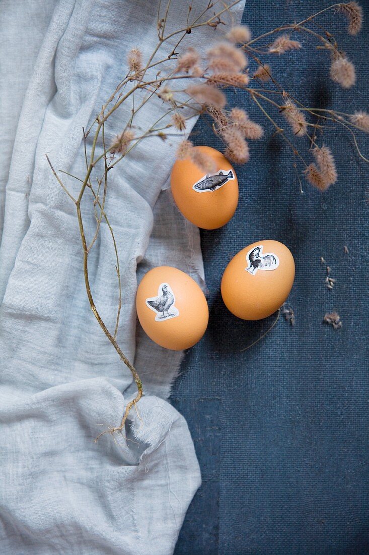 Ostereier mit aufgeklebten Tiermotiven und Trockenzweig auf blauem Stoff