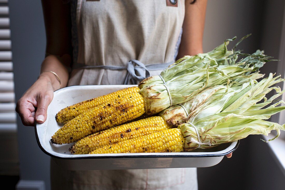 A woman holding an enamel dish of grilled corn on the cob