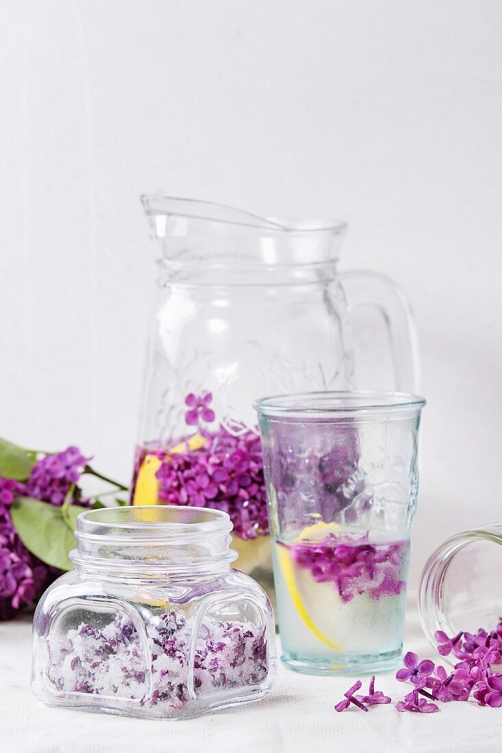 Glass jar of lilac flowers in sugar, glass and pitcher of lilac water with lemon and branch of fresh lilac