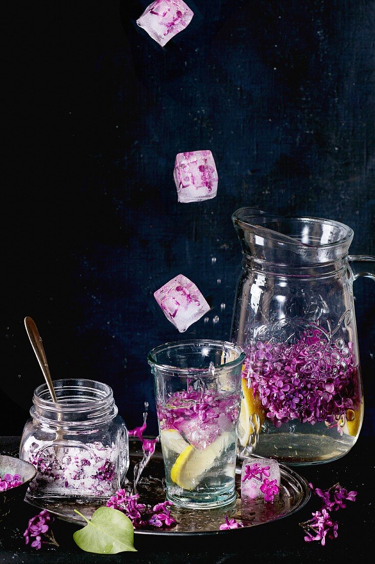 Ice cubes with flowers falling into glass of lilac lemonade with lemon