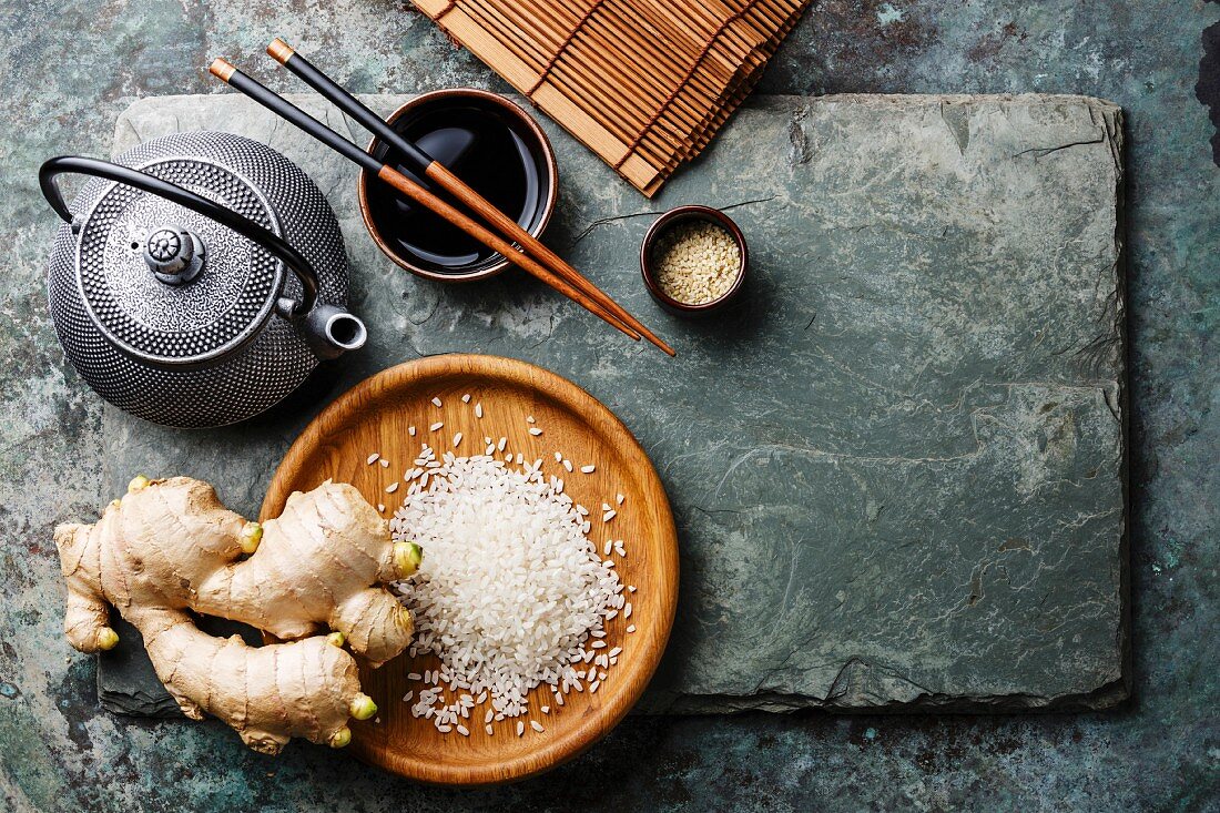 Raw white rice, green tea teapot, soy sauce and sushi chopsticks on gray stone slate background