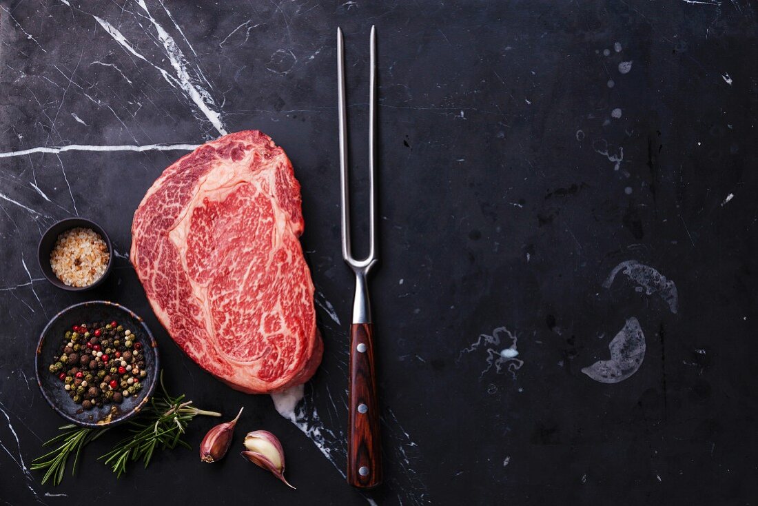 Rohes Black Angus Ribeye Steak mit Fleischgabel und Gewürzen auf dunklem Untergrund
