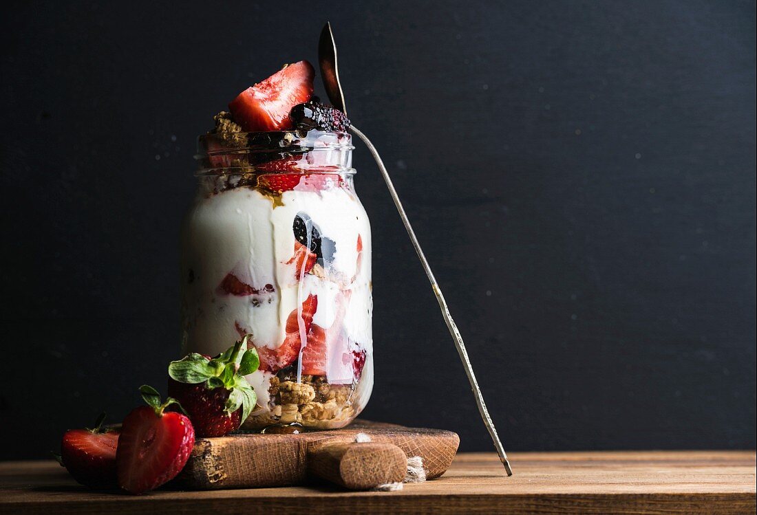 Hafermüsli mit Joghurt, Erdbeeren, Maulbeeren, Honig und Minze in Glas vor schwarzem Hintergrund