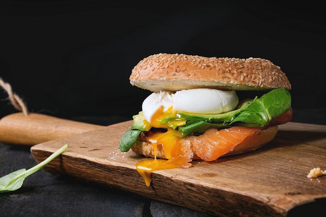Bagel with salted salmon, spinach, avocado and soft boiled egg with liquid yolk on wooden chopping board