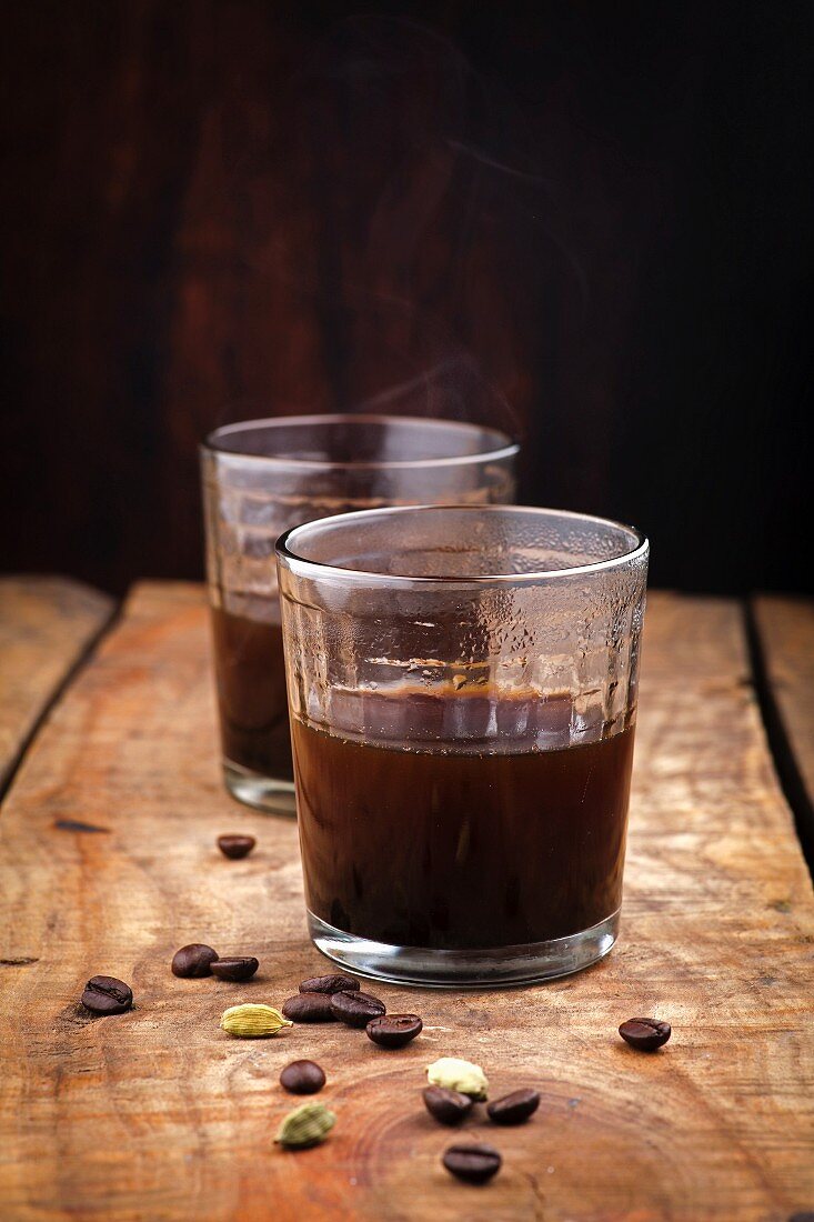 Kaffee in zwei Gläsern, daneben Kaffeebohnen und Kardamom