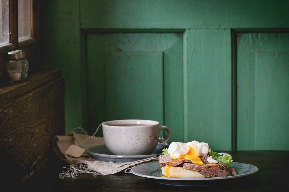 Frühstück mit Fleisch-Sandwich, pochiertem Ei und einer Tasse Tee