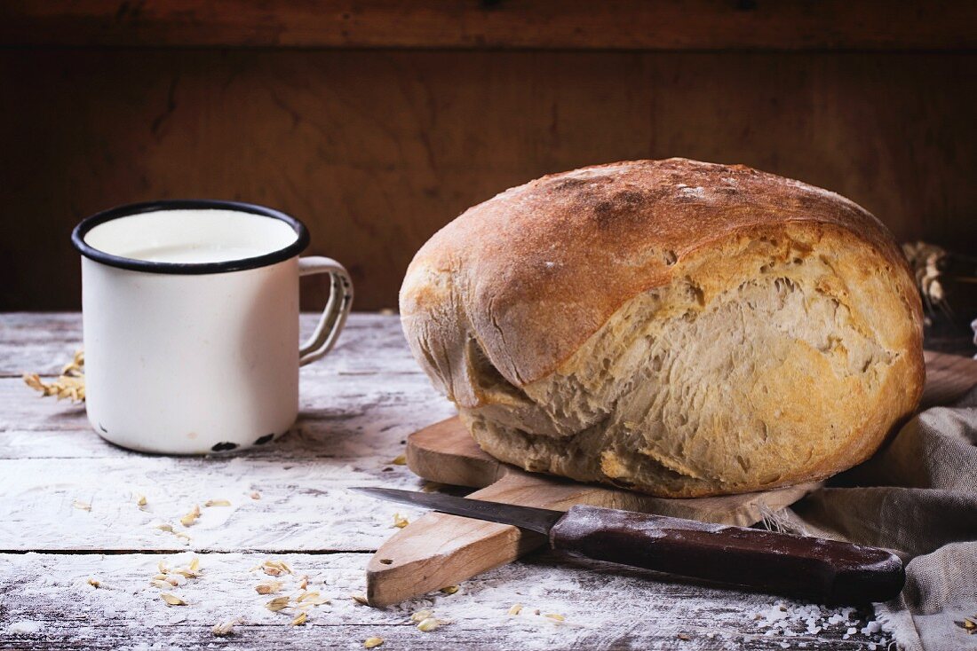 Fresh homemade bread on wooden cutting board with vintage knife and mug of milk