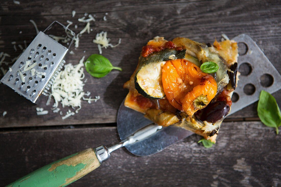 A slice of homemade pizza with pepper, courgette, olive and basil and a cheese grater