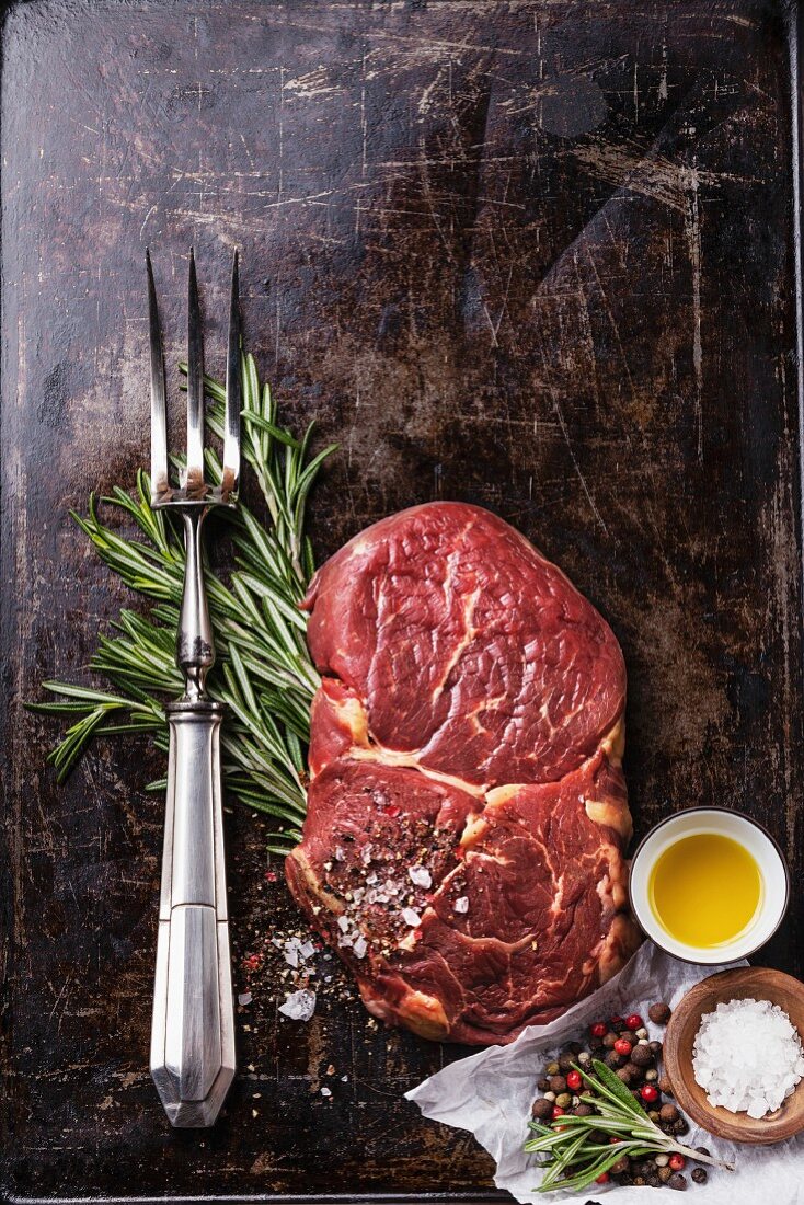 Raw fresh meat Ribeye Steak, seasoning and meat fork on dark background