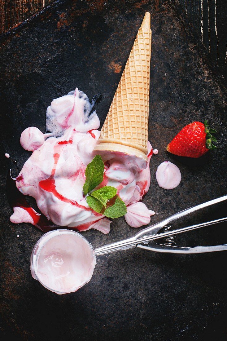 Wafer cone with strawberry ice cream with fresh strawberries, mint and metal spoon over black table