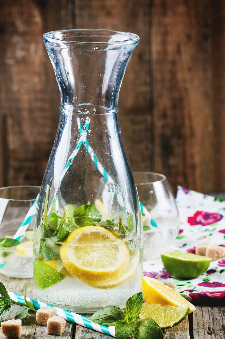 Glass pitcher of homemade lemonade with lemon, lime, sugar and mint on old wooden table