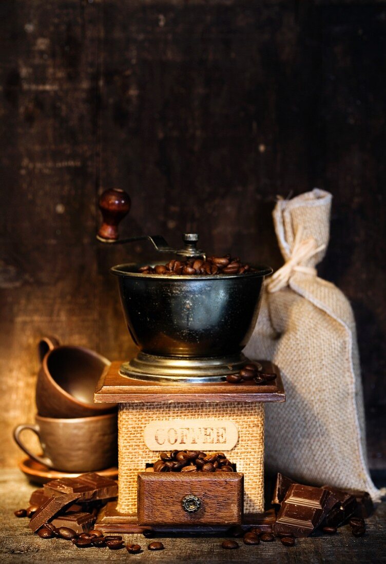Stiillleben mit antiker Kaffeemühle, Kaffeesack, Kaffeetassen und Schokolade auf rustikalem Tisch