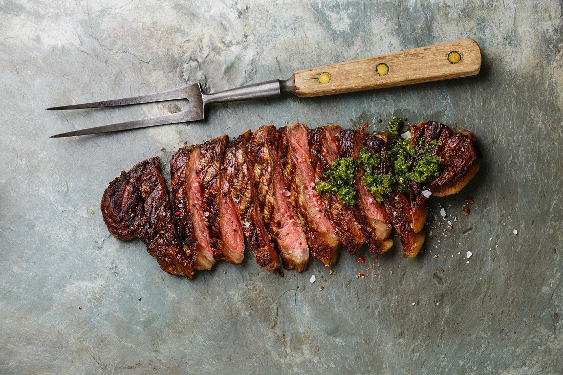 Gegrilltes Striploin Steak in Scheiben mit Chimichurri-Sauce und Fleischgabel auf grauem Steinhintergrund