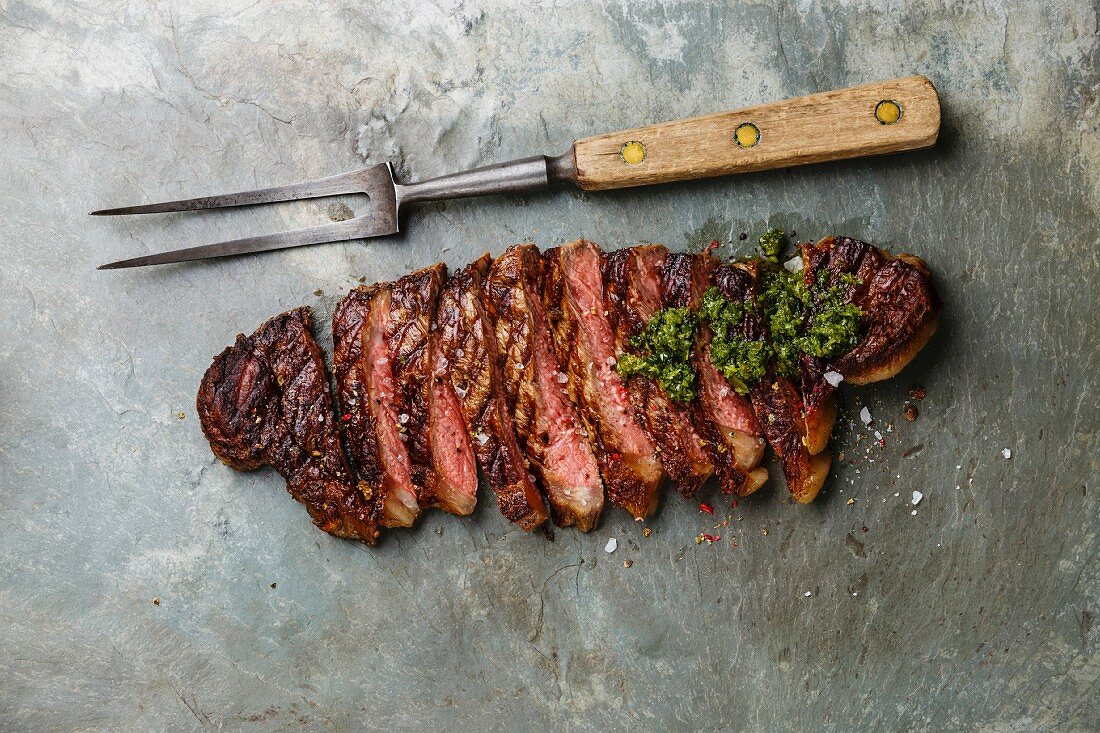 Sliced grilled beef barbecue Striploin steak with chimichurri sauce and meat fork on gray stone slate background