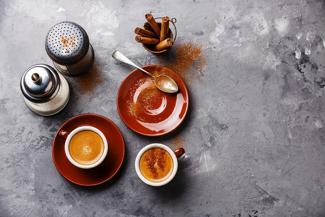 Kaffee in Tassen mit Zimt und Zuckerstreuer auf grauem Untergrund