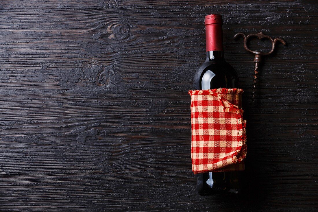 Weinflasche in kariertes Tuch gewickelt neben Korkenzieher auf schwarzem Holzuntergrund